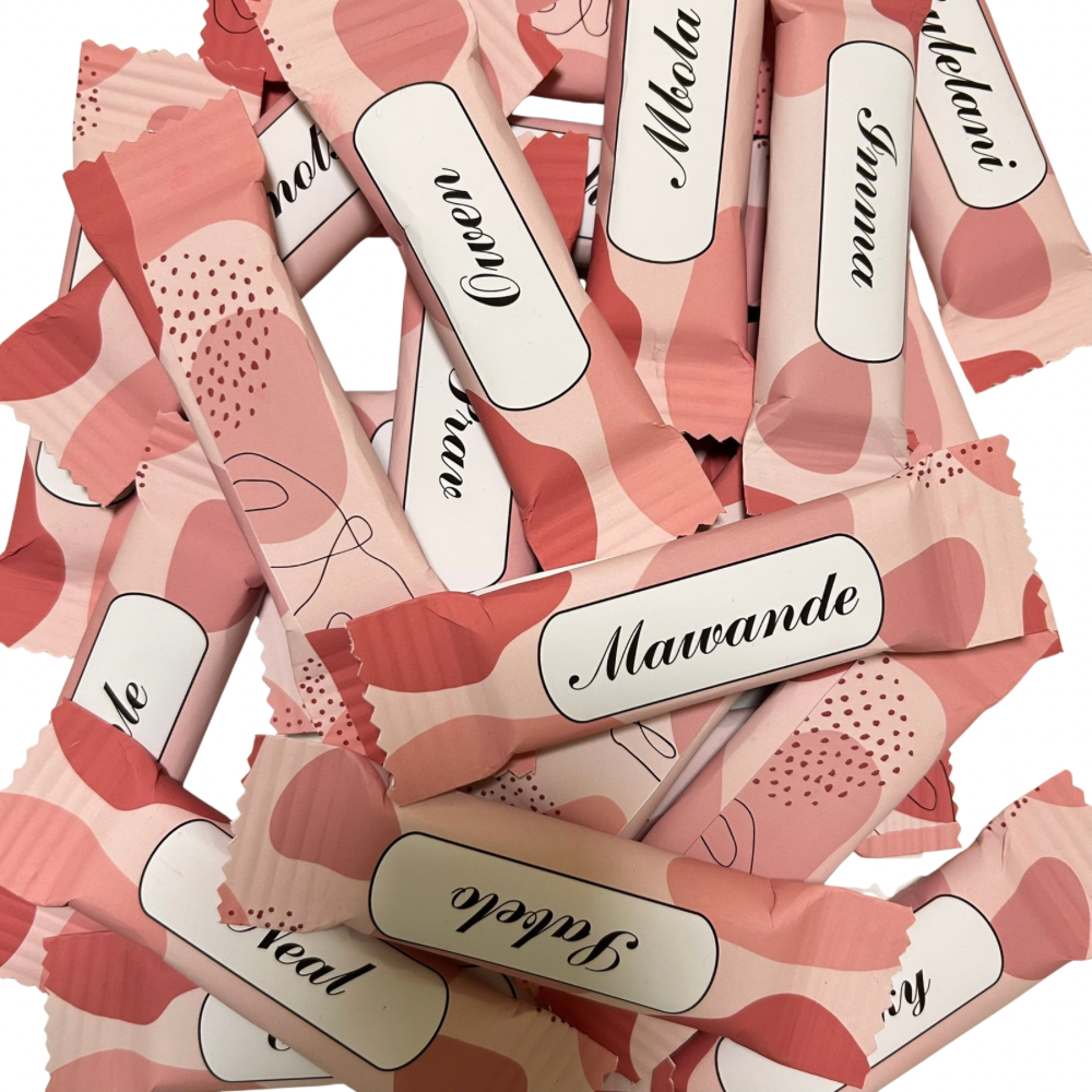 A pile of individually wrapped chocolates on a plain white background. Each wrapper is light pink with a darker pink abstract pattern and a white label with black script text with names.